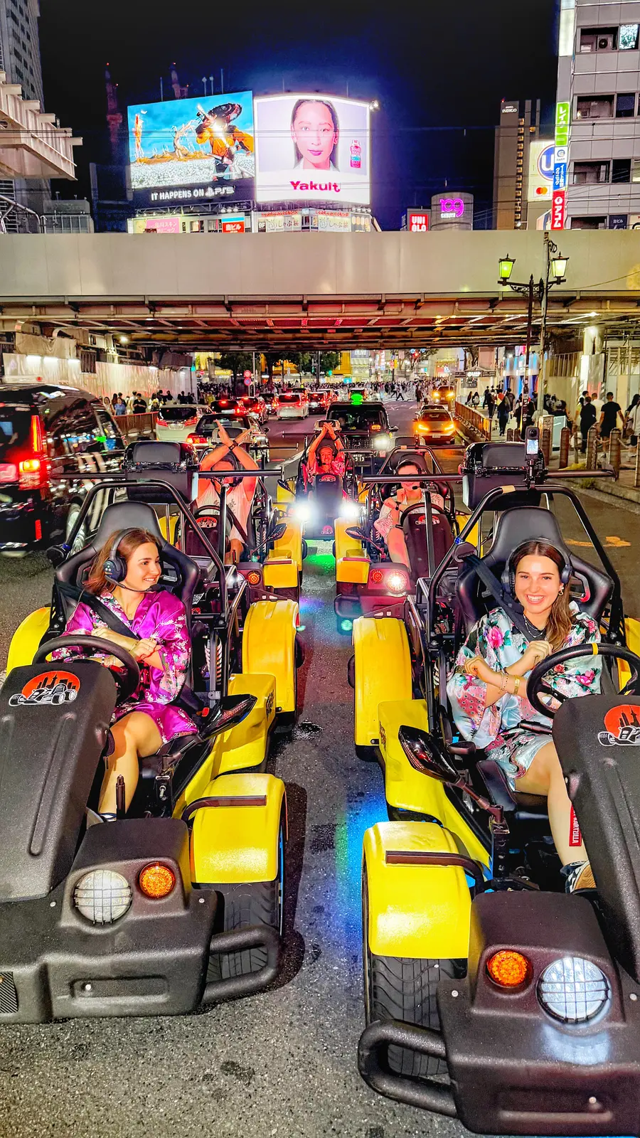 Convoy of go-karts parked under a rail bridge in Shibuya at night, riders in colorful kimono-style robes, beneath the iconic Yakult and Apple TV+ billboards.