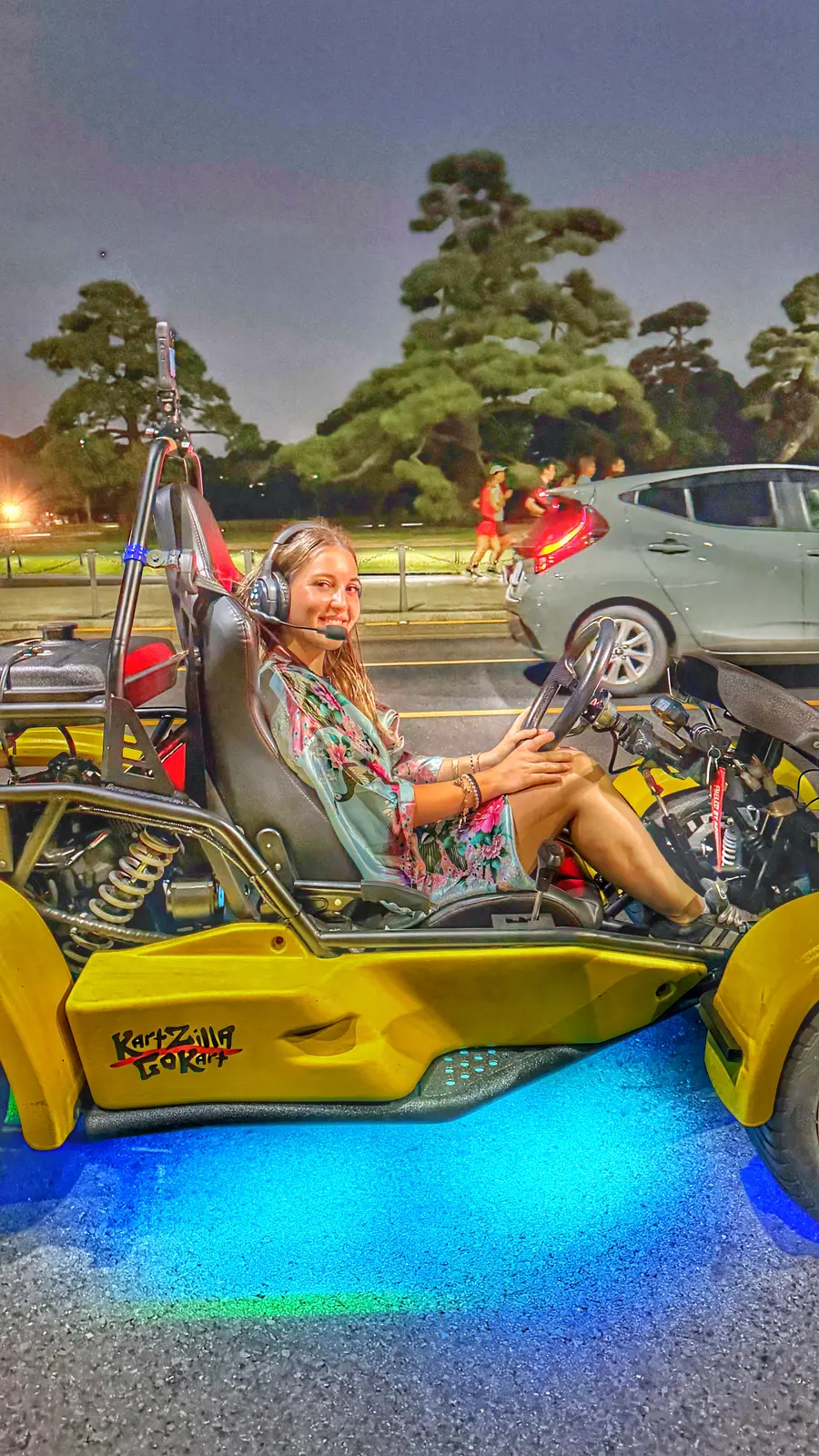 Driver in a teal floral kimono-style robe seated in a KartZilla go-kart with vivid blue underglow, cruising past the Imperial Palace moat at dusk.